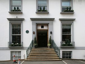 A flight of stone steps leads from an asphalt car park up to the main entrance of a white two-story building. The ground floor has two sash windows, the first floor has three shorter sash windows. Two more windows are visible at basement level. The decorative stonework around the doors and windows is painted grey.